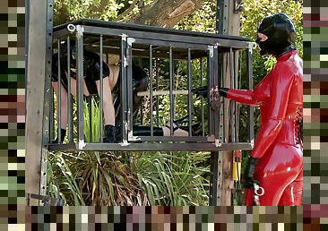Latex slave girl in a cage and her mistress play outdoors